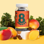 A jar of 8Greens Super Greens Gummies (QVC) for immunity boost is set among kale and mangoes on a light backdrop. The label has red and yellow hues with nutritional details, while scattered gummies surround the jar.