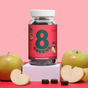 A jar of 8Greens Super Greens Gummies (QVC) is set against a pink backdrop, surrounded by whole and sliced green apples. Apple-flavored for immune support, the gummies label prominently features a green 8.