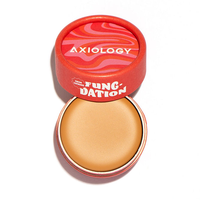 A round, peach-toned Fungdation - Mushroom Foundation by Axiology sits in an open red container labeled with the brand and product name, its colorful, wavy lid placed beside it.