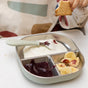 A child’s hand holds a biscuit above a divided plate with yogurt, purple fruit puree, and banana slices. A small spoon from the BEABA Stainless Steel Toddler Cutlery Set rests in the yogurt section.