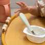 A baby’s hand stretches toward a cup of yogurt with a spoon from the BEABA Stainless Steel Toddler Cutlery Set resting inside, all placed on a wooden tray beside a beaded string and a pink sippy cup. Only the baby’s hand and part of their arm are visible as they eagerly reach for the ergonomic kids utensils crafted for little hands.