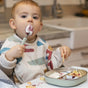 A toddler in a bib sits at a table, happily eating with a BEABA Stainless Steel Toddler Cutlery Set and holding food. With food smeared on their face, the child appears to be enjoying their meal in a kitchen setting, making mealtime even more delightful with the help of this eco-friendly cutlery set.