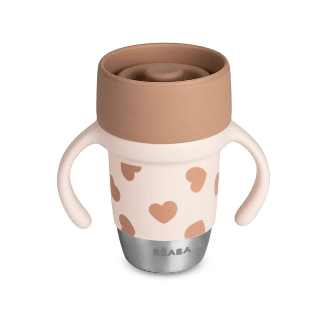 Introducing the BEABA Stainless Steel Learning Cup, Click & Drink: a beige and pink cup featuring two easy-grip handles, brown heart patterns, a brown lid, and a sturdy silver base labeled BÉABA. Designed with a 360° valve-free, spill-proof system for effortless sipping and mess-free learning.