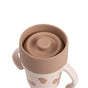 A BEABA Stainless Steel Learning Cup, Click & Drink, in beige and light brown with two handles, a spill-proof lid, and heart-shaped decorations on the body, displayed against a white background.