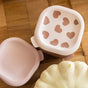 Two light pink, square BEABA Stainless Steel Bowls with silicone lids featuring heart-shaped cutouts are placed on a wooden surface next to a pale gourd or squash, offering a stylish and practical solution for baby food storage. These microwave-safe bowls provide both convenience and durability for modern families.
