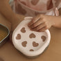 A child holds a pink silicone lid with heart-shaped cutouts over a BEABA Stainless Steel Bowls with Lid Set (Microwave-Safe) on a wooden table.