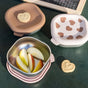 Heart-shaped cookie and apple slices in a metal bowl, alongside two BEABA Stainless Steel Bowls with Lid Set—one featuring a heart-patterned silicone cover and the other with a solid lid—rest on a green surface, perfect for keeping snacks fresh in these microwave-safe baby food containers.