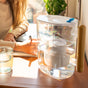 A person sits at a table writing in a notebook. On the table are a glass of water and the Epic Water Filters USA Pure XP Water Filter Pitcher, NSF certified and designed to reduce fluoride, PFAS, and parasites, with sunlight streaming in through a nearby window.