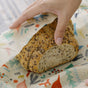 A hand wraps a seeded loaf of bread in the Bees Wrap Bread Wrap - Holiday, an extra large, reusable beeswax wrap with colorful festive designs, helping to reduce single-use waste.