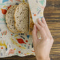 A hand wraps slices of bread in the Bees Wrap Bread Wrap - Holiday on a wooden surface, keeping loaves fresh and cutting down on single-use waste.