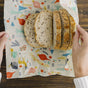 Hands wrapping slices of multigrain bread in the Bees Wrap Bread Wrap - Holiday, a colorful, nature-themed beeswax food wrap on a wooden table—ideal for reducing single-use waste.