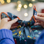 Close-up of hands tying Bees Wrap Reusable Gift Wrap, made from organic cotton with a festive pattern. Warm, blurred lights create a cozy holiday atmosphere in the background.