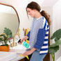 A woman in a blue and white striped shirt cleans a bathroom sink with Aunt Fannies All-Purpose Cleaner and a yellow cloth. Nearby are towels, a potted plant, and a round mirror above the sink.