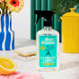 A bottle of Aunt Fannies All-Purpose Cleaner rests on a counter by a sponge, half a lemon, a yellow container, and a floral vase, with a green curtain and colorful decor in the background.