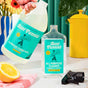 A hand pours Aunt Fannies All-Purpose Cleaner, Refill into a spray bottle on a kitchen counter with a sliced lemon, yellow canister, blue and white vase with flowers, and a spray nozzle nearby.