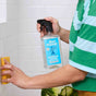 A person in a green striped shirt uses a yellow sponge and holds Aunt Fannies Bathroom Cleaner spray while cleaning white tiled bathroom walls.