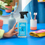 A hand holds a bottle of Aunt Fannies Bathroom Cleaner on a blue counter, next to a folded yellow towel, a toothbrush holder with two toothbrushes, a soap dispenser, and potted plants.