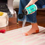 Wearing blue jeans and orange-checkered socks, a person sprinkles Aunt Fannies Carpet Refresher from a teal container onto a pink and white striped rug in the living room.