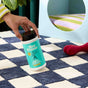 A hand sprinkles Aunt Fannies Carpet Refresher, a natural odor remover, onto a blue and white checkered carpet. Nearby are a red dog toy and a striped-cushion pet bed.