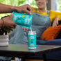 A person pours Aunt Fannies Carpet Refresher - Refill into a container on a table, while someone in a yellow shirt and overalls sits on a blue couch in the background.