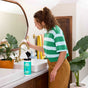 A woman in a green and white striped shirt wipes a gold faucet with Aunt Fannies Cleaning Vinegar Wipes in a bright bathroom. Plants decorate the counter, and colorful towels hang in the background.