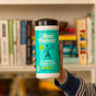 A hand holds a container of Aunt Fannies Cleaning Vinegar Wipes with a turquoise label highlighting eucalyptus scent and vinegar-powered cleaning, in front of a bookshelf.