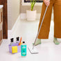 Wearing green socks and brown pants, a person mops a white tile floor with Aunt Fannies Multi-Surface Floor Cleaner. Nearby is a caddy with spray bottles, yellow cloth, scrub brush, cleaner, and a potted plant in the background.