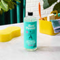 A bottle of Aunt Fannies Multi-Surface Floor Cleaner sits on a white countertop beside a yellow sponge and cleaning caddy, with green plants and a white brick wall in the background.