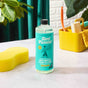 Aunt Fannies Multi-Surface Floor Cleaner sits on a light countertop next to a yellow sponge. Cleaning supplies in a yellow container, lush plants, and a green curtain are visible in the background.