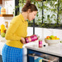 A woman in a yellow sweater pours Aunt Fannies FlyPunch! Fruit Fly Trap Refill into a glass in a bright kitchen with fruit and green plants, demonstrating an easy way to reduce plastic waste.