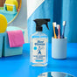 A bottle of Aunt Fannies Glass & Window Cleaner sits on a blue surface near a cup with three toothbrushes, a pink towel, yellow sponge, and a blue storage basket in a bright, colorful bathroom setting.