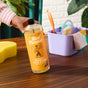 A hand holds a bottle of Aunt Fannies Hardwood Floor Cleaner on a wooden table, with a yellow sponge and a purple cleaning caddy containing cloths and a scrub brush in the background.
