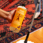 A hand holds a bottle of Aunt Fannies Hardwood Floor Cleaner over a red and blue patterned rug, with a mop and part of a leg in the background.