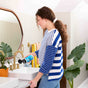 A woman in a blue and white striped shirt pours Aunt Fannies Sink Refresher into a bathroom sink with gold fixtures. Green plants and patterned wallpaper add decor to the countertop.
