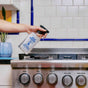 A hand holds a spray bottle labeled Aunt Fannies Stainless Steel Cleaner, aimed at a stove with control knobs in a kitchen with white tiled walls and a potted plant.