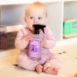 A baby in a pink outfit sits on the floor, putting the spray nozzle of Aunt Fannies Toy + Highchair Cleaner in their mouth. Shelves and toys can be seen in the background.