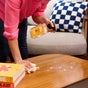 Wearing a pink shirt, a person cleans a wooden coffee table with Aunt Fannies Wood Surface Cleaner and a cloth. Soap bubbles appear on the table, while books and a checkered pillow are visible in the background.
