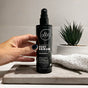 A hand holds a black bottle labeled All Good Body Serum on a tiled countertop, featuring organic ingredients. In the background sits a small green succulent and a rolled-up white towel.