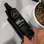 A hand holds a black bottle of All Good Body Serum, infused with Organic Moringa Oil and grapefruit scent. The label is in English and French. Nearby, a white planter with greenery peeks into view on the pristine white surface.