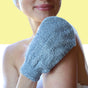 A person uses the Afterspa Exfoliating Bamboo Mitt on their shoulder, smiling softly. The background is split yellow and white, with their face partially visible.