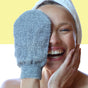 A smiling person with a towel on their head holds the Afterspa Exfoliating Bamboo Mitt made from natural bamboo fibers over one eye, while touching their face, set against a yellow and white background.