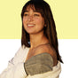 A woman with straight brown hair and bangs smiles, wearing an off-the-shoulder white top. She holds the Afterspa Exfoliating Bamboo Mitt, made from natural bamboo fibers, over her shoulder against a yellow and white background.