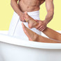 A person uses the Afterspa Foot Brush to massage someone’s feet as they relax in a white bathtub, both wrapped in white towels, set against a light yellow background.