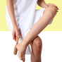 A person in a white towel uses the Afterspa Foot Brush to gently brush the bottom of their foot, with one leg crossed over the other against a yellow and white background that evokes a relaxing shower spa experience.