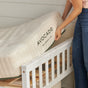 A person lifts an Avocado Green Mattress Eco Organic Kids Mattress onto a white crib frame, highlighting the label of this organic hybrid mattress.