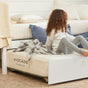 A child with curly hair sits on an Avocado Green Mattress Eco Organic Kids Mattress with a blanket, next to another bed with pillows in a bright, cozy bedroom.