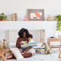 In a cozy nursery with toys, plants, and animal-themed art, a woman reads to a child sitting on the floor. An Avocado Toddler Pillow rests nearby, along with a crib and stuffed dog.