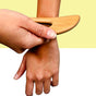 A hand uses the Afterspa Body Gua Sha, a wooden massage tool with a curved edge, on another persons inner forearm against a yellow and white backdrop.