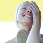 A smiling woman with a towel wrapped around her head gently exfoliates her neck using the Afterspa Cleansing Sisal Mitt, enjoying self-care against a split yellow and white background.