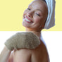 A smiling woman with a white towel wrapped around her head uses the Afterspa Cleansing Sisal Mitt on her shoulder, enjoying gentle exfoliation against a yellow and white background.